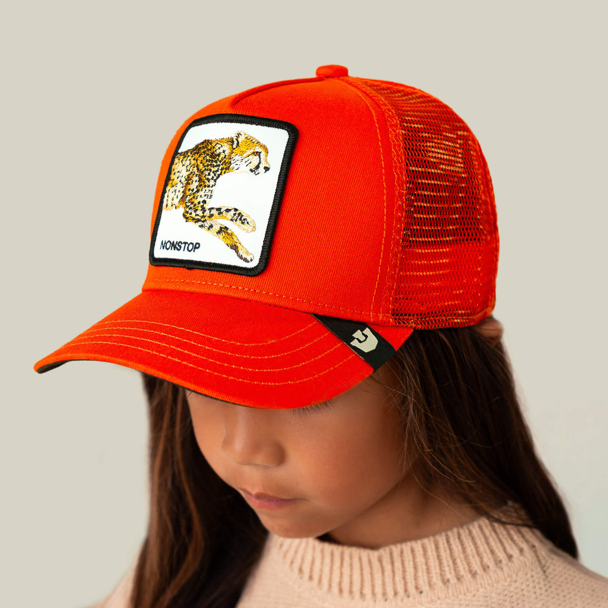 A child wearing Goorin Bros.’ The Nonstop Cheetah Mini Noir kids cap, which features an orange crown and a patch with a cheetah and the word “NONSTOP.” The child is looking down and dressed in a light-colored knit sweater.