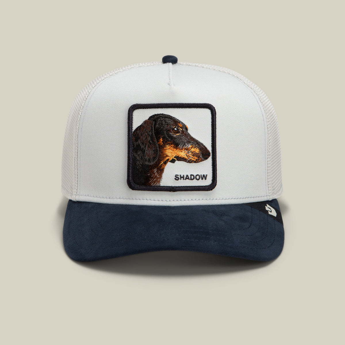 The Suede Shadow by Goorin Bros. is a white and navy trucker hat featuring a suede brim and a patch with an embroidered dachshund profile and the word “SHADOW” below—an essential dog patch hat for canine enthusiasts.