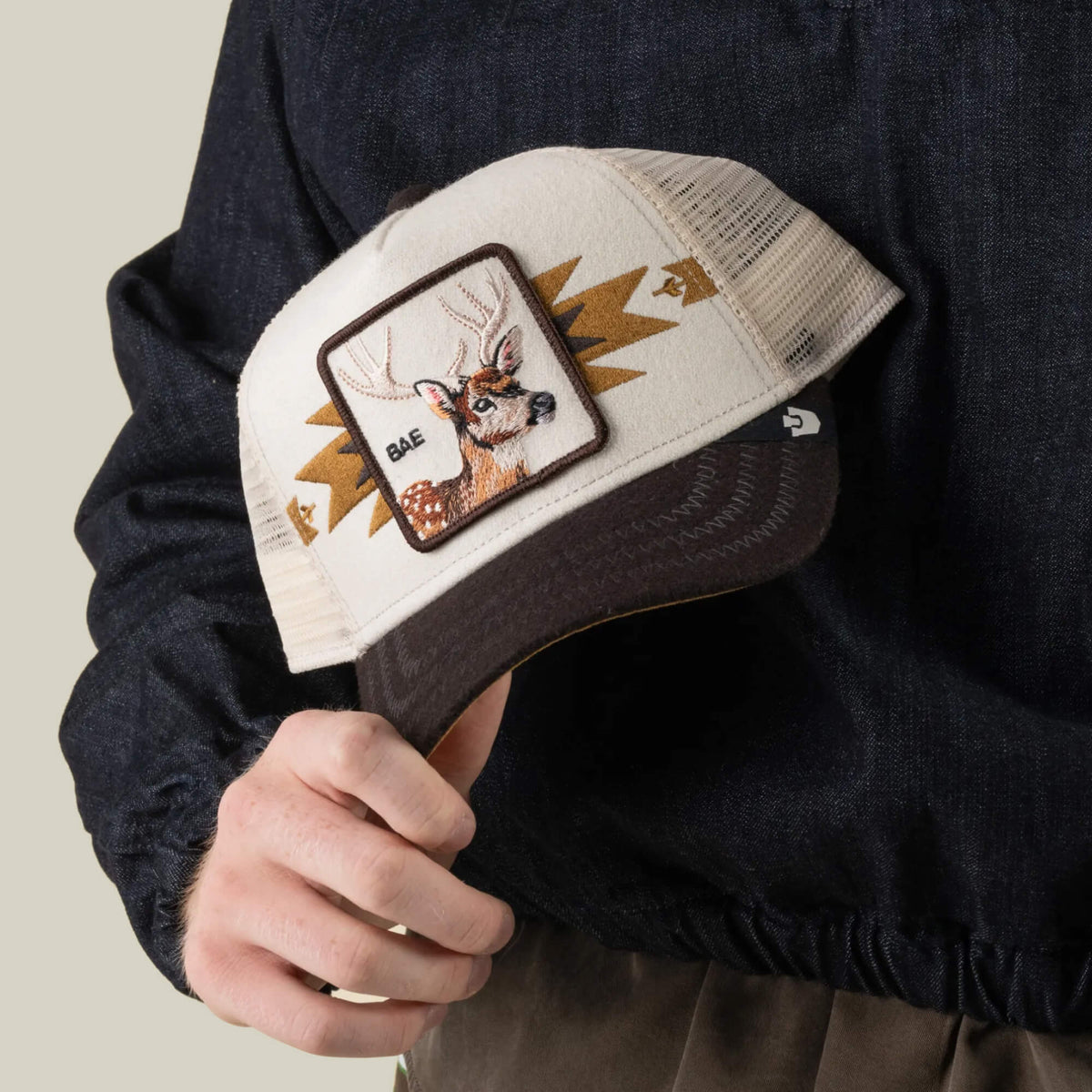 A person holds the Goorin Bros. Canyon Bae trucker cap—featuring a beige and brown colorway, a mesh back in a premium blend, and a patch with a deer’s head and the word “BAE”—paired stylishly with a dark jacket.