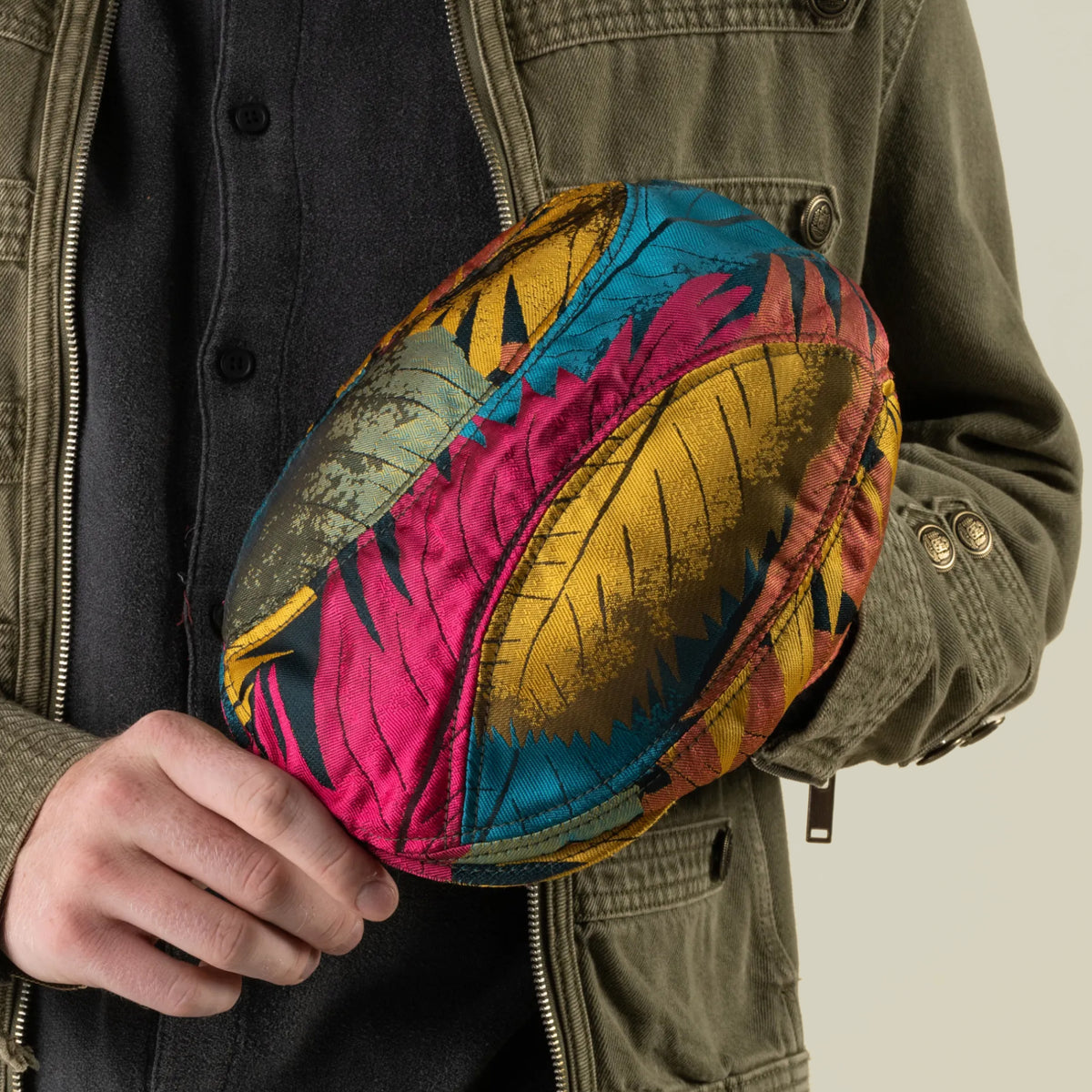 A person in a green jacket holds a colorful, patterned rugby ball adorned with yellow, pink, and blue feather-like designs. Their look is further elevated by the Goorin Bros. Upland Avenue multi-color headwear, which brings even more vibrant flair to the ensemble.