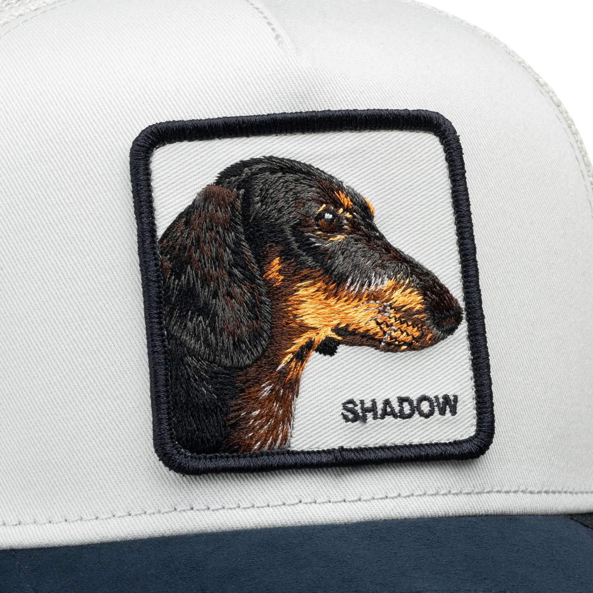 Here’s a rewritten sentence incorporating the provided product data: Close-up of the Goorin Bros. Suede Shadow trucker hat, featuring a suede brim and a stitched patch with a dachshund’s head in profile above the embroidered word “SHADOW.”.