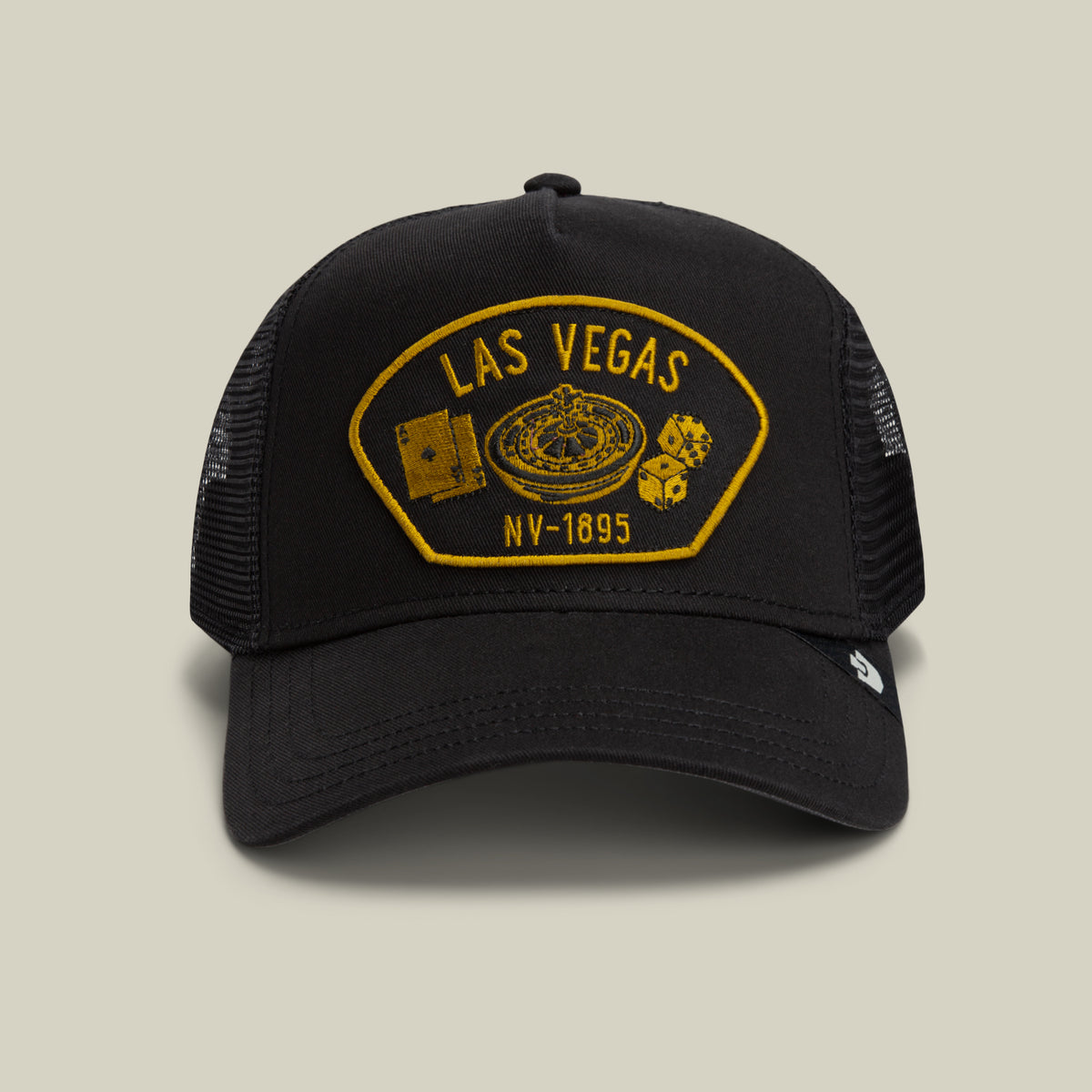 The Goorin Bros. “Las Vegas” trucker hat features a black design with a gold embroidered patch displaying playing cards, a roulette wheel, and dice, set against a plain light gray background. Its classic mesh back ensures breathability and comfort.