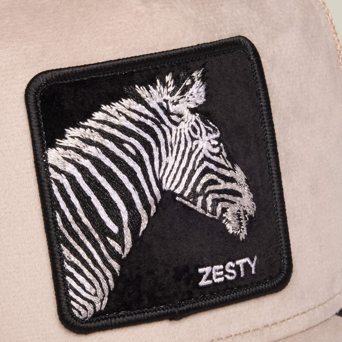 Square patch with an embroidered zebra head featuring monochrome stripes and the word “ZESTY” stitched below, sewn onto a beige velour fabric background—a unique detail from Goorin Bros.’ Velour Zesty, perfect for any zebra trucker hat.