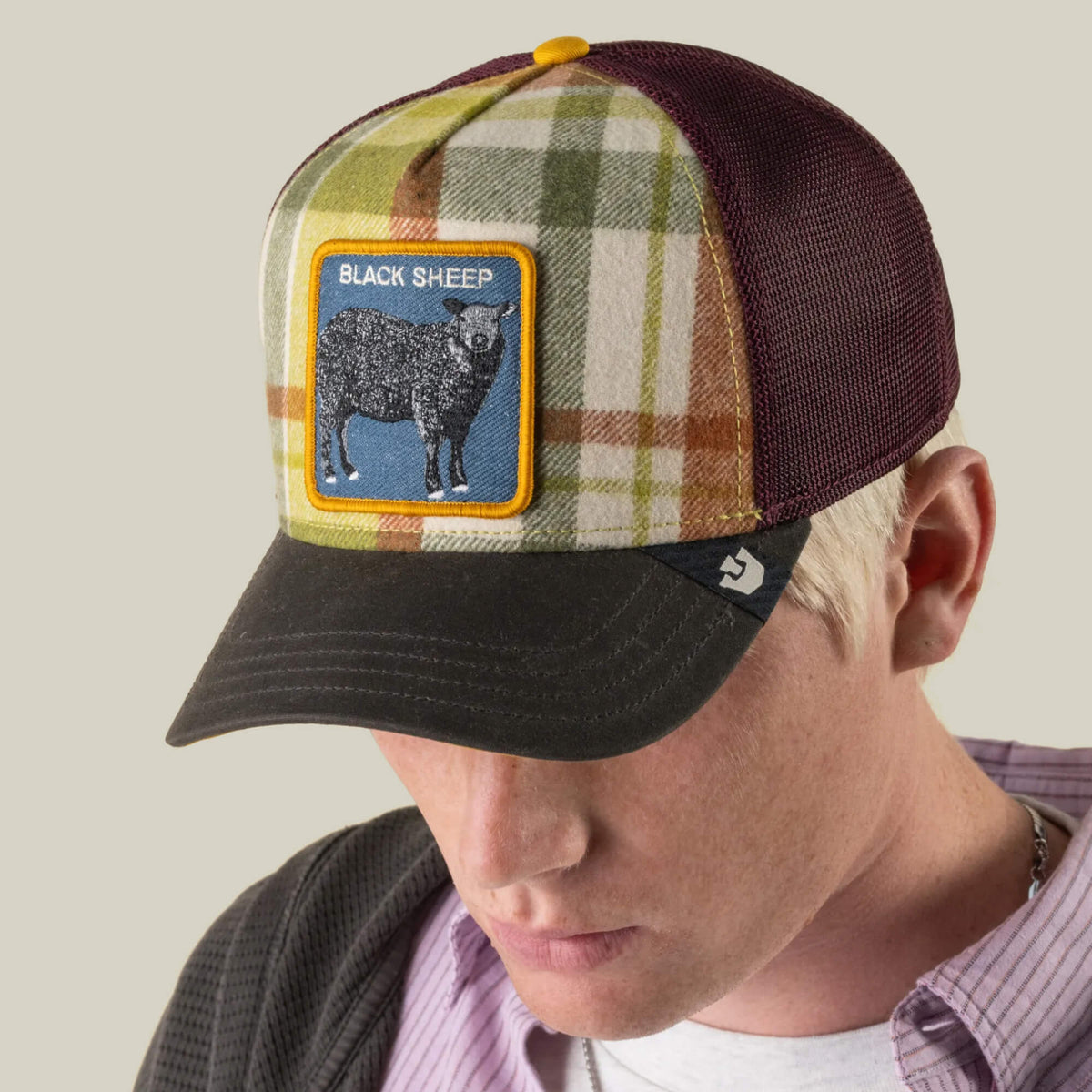 A person wearing the Goorin Bros. “Brit Sheep” fall plaid trucker hat with a mesh back and a BLACK SHEEP patch, looking downward against a plain background.