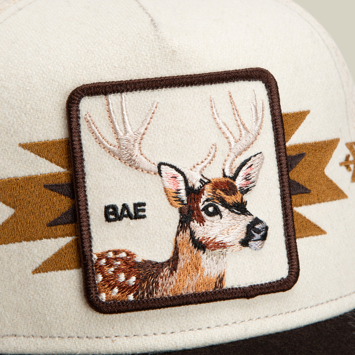 The Goorin Bros. Canyon Bae cap features a cream-colored mesh back and brown brim, highlighted by an embroidered patch of a deer with antlers and “BAE” stitched beside it. Geometric brown accents complete this premium blend antler-themed hat.