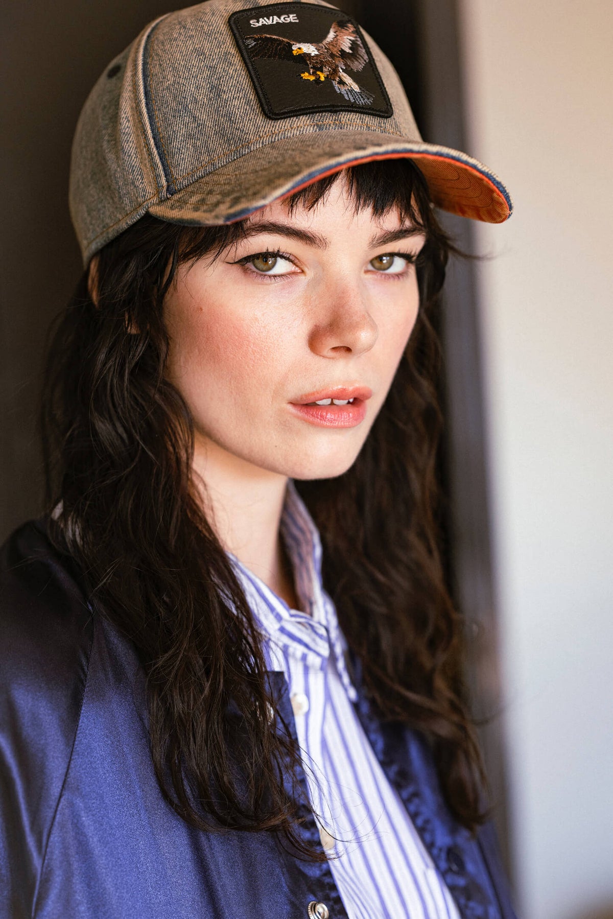 A woman with long dark hair wears a Goorin denim cap with an eagle patch, a blue striped shirt, and a satin jacket, looking at the camera in natural light.