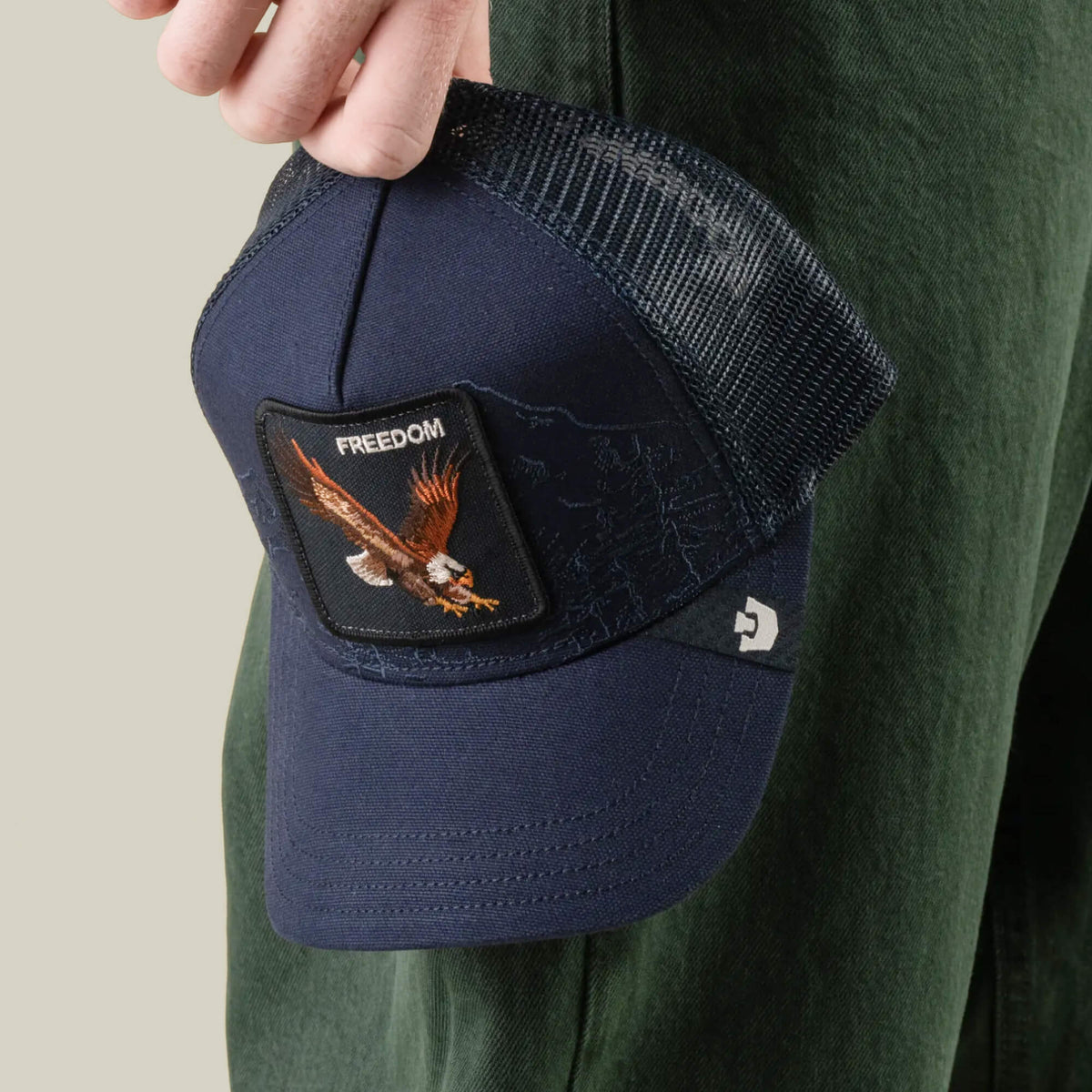 A person holds the Goorin Bros. Badlands Eagle navy trucker hat, featuring an eagle patch labeled “FREEDOM” and a breathable mesh back, which stands out against dark green pants.