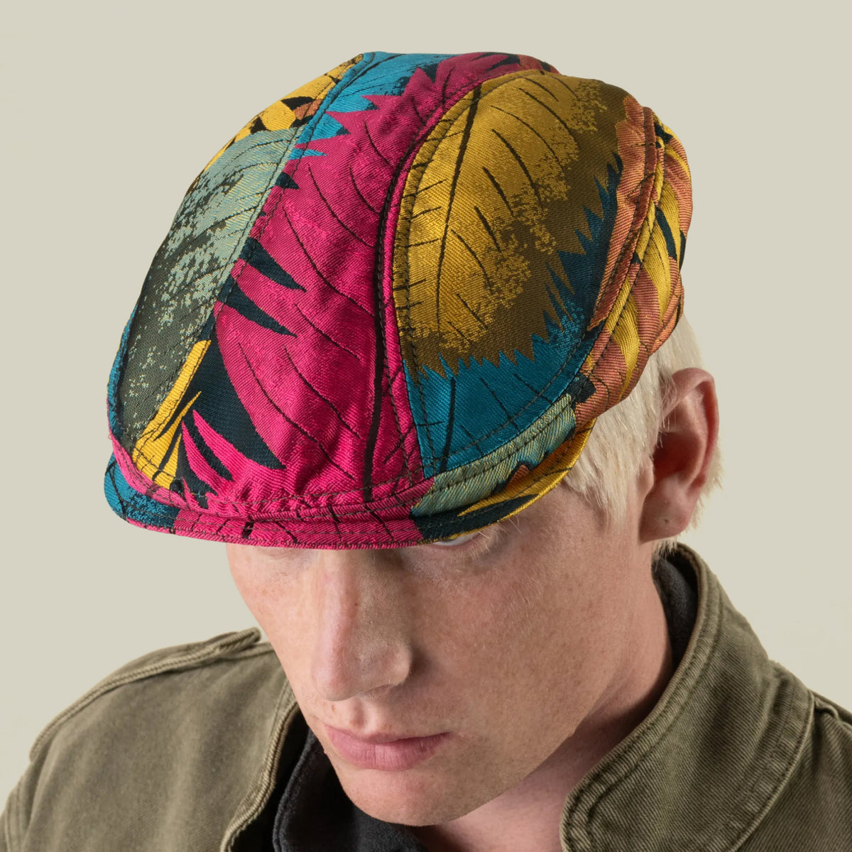 A person with light hair wears the Upland Avenue multi-color, leaf-patterned headwear by Goorin Bros., hand-cut in the USA, paired with an olive jacket, against a plain background.
