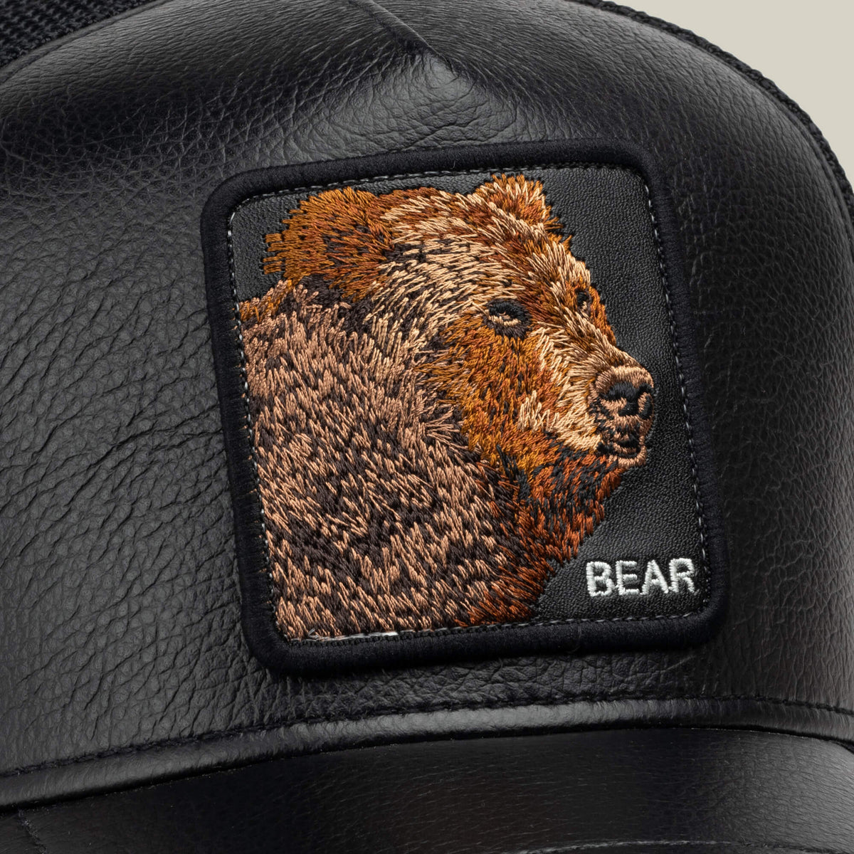 The Goorin Bros. Faux Leather Bear is a black trucker cap with breathable mesh sides, featuring a rectangular embroidered patch of a brown grizzly bear and the word “BEAR” stitched in white in the bottom right corner.