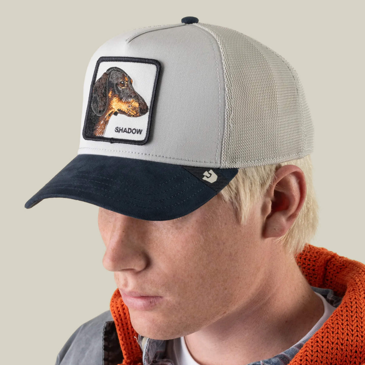 A person wearing the Goorin Bros. Suede Shadow trucker hat—a light gray mesh cap with a navy suede brim and a dachshund patch labeled “SHADOW”—paired with a gray shirt and an orange-lined jacket.