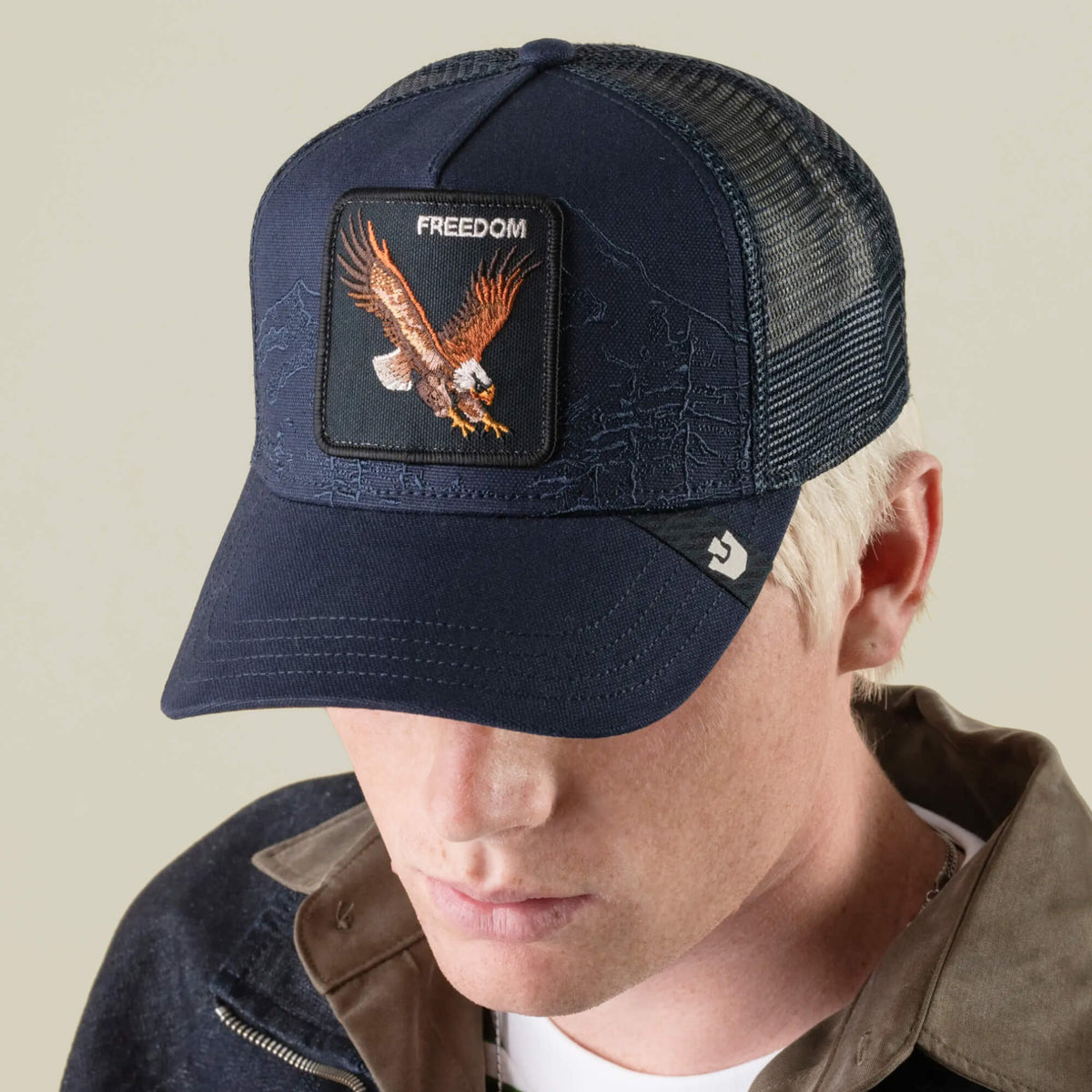 A person with light hair is looking down while wearing the Goorin Bros. Badlands Eagle navy blue trucker hat, which features a breathable mesh back and an eagle patch labeled “FREEDOM.” The individual is dressed in a dark jacket layered over a beige top.