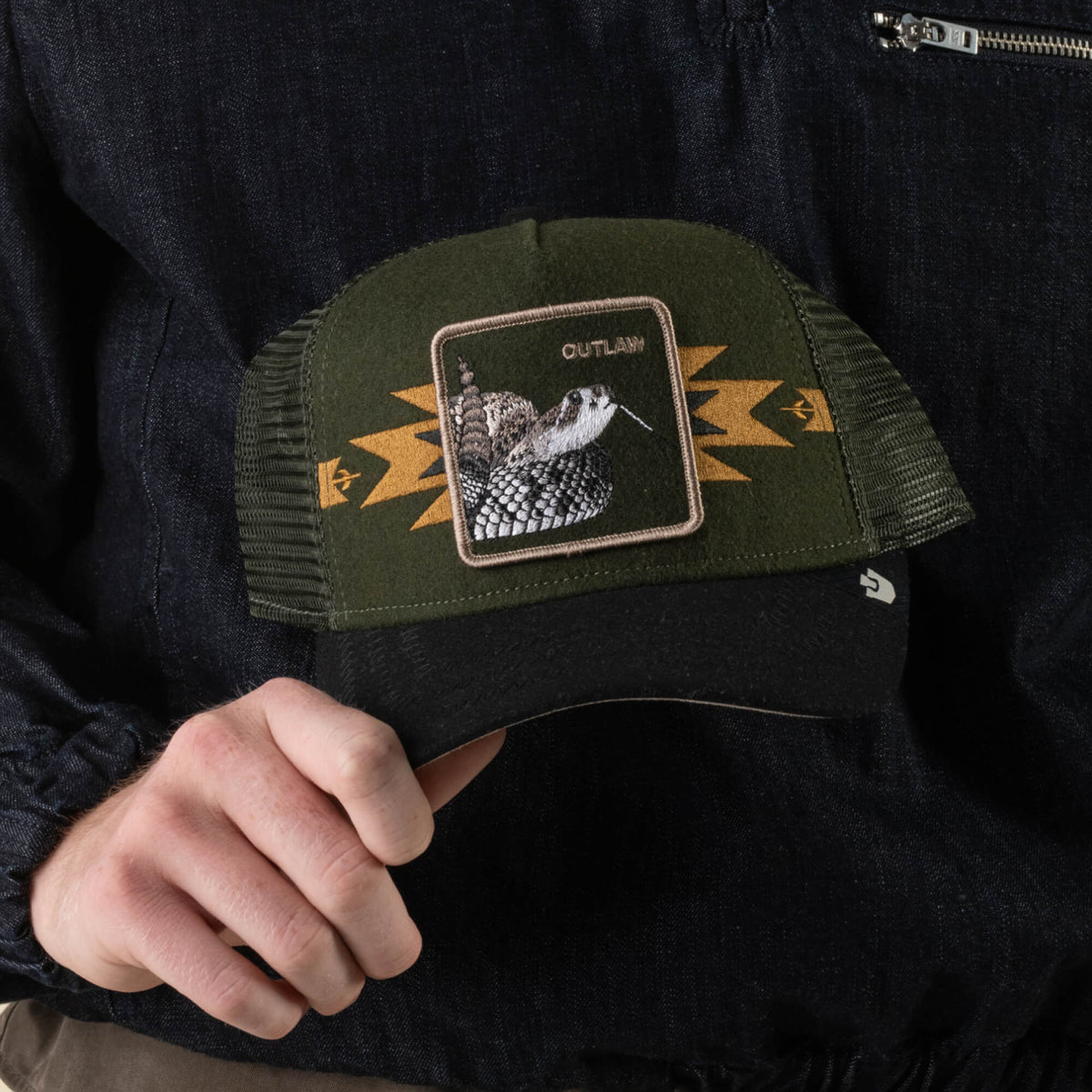 A person wears the Goorin Bros. Canyon Outlaw hat, a green mesh wool blend trucker featuring a rattlesnake patch labeled “OUTLAW,” channeling undeniable outlaw energy. The look is completed with a dark jacket.