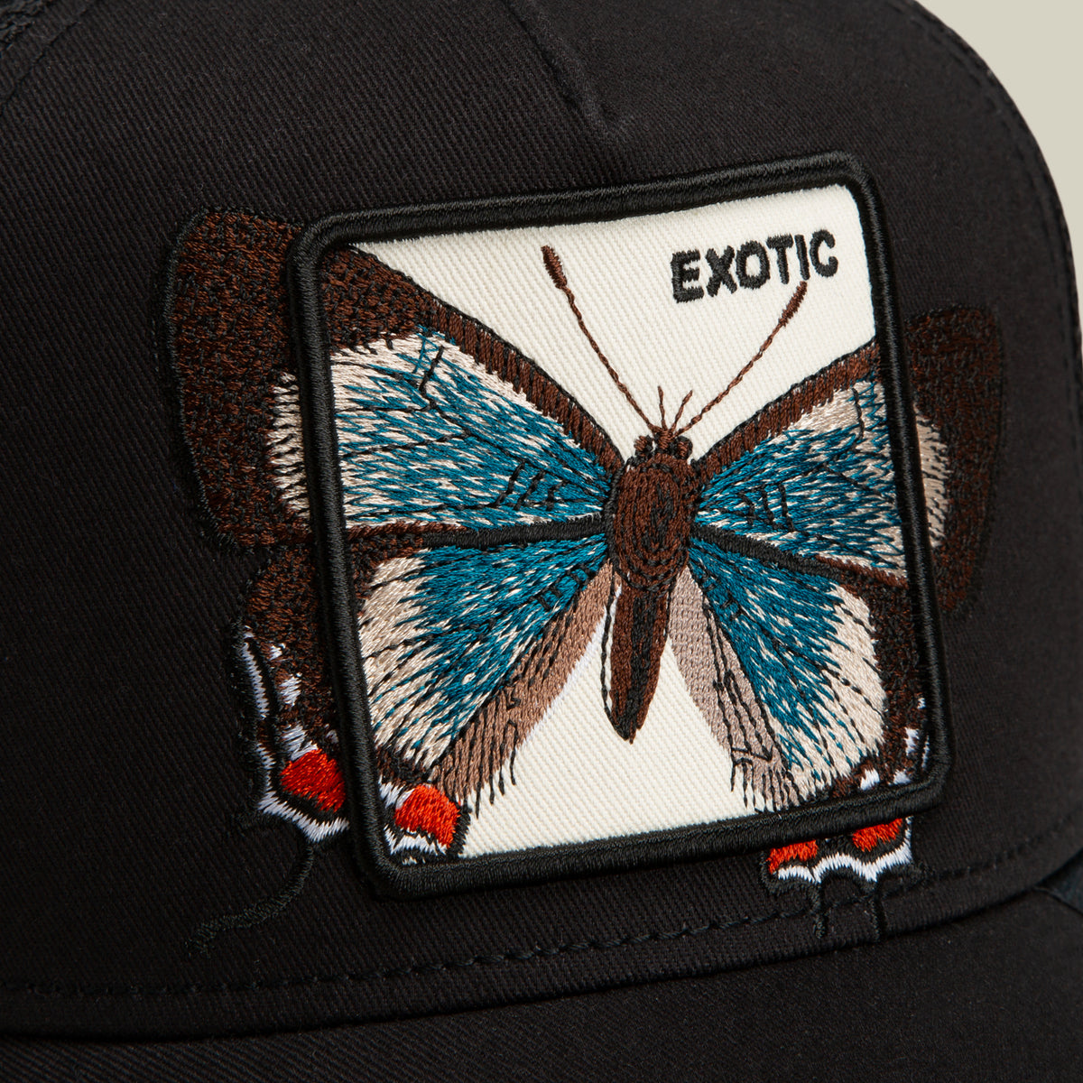 The Goorin Bros. Over the Line Butterfly is a black, breathable mesh cap featuring a rectangular patch with an embroidered blue and brown butterfly and the word “EXOTIC” in black letters on a white background.