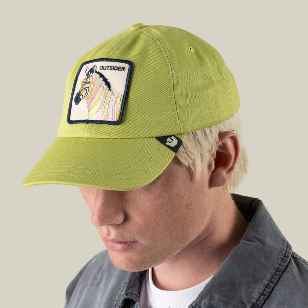 A person with light blond hair wearing a light green Goorin Bros. Outsiderland Papa Cap baseball cap featuring a zebra patch labeled “OUTSIDER,” along with a gray jacket, faces slightly downward against a plain background; the handmade cap is proudly made in the USA.