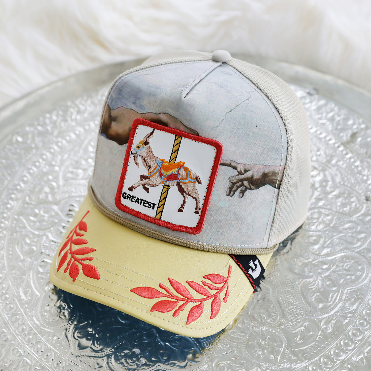 A The Farm “Maximum” mesh cap, featuring a printed mural of hands, a “Greatest” goat patch, and red leaf embroidery on the yellow brim, rests on a patterned silver tray. *Please note: All Sales Are Final.*.