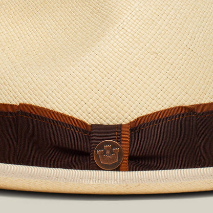 Close-up of the Hartman straw hat by Goorin Bros., featuring a brown and orange grosgrain band, contrast edge binding, and a round metal castle emblem.