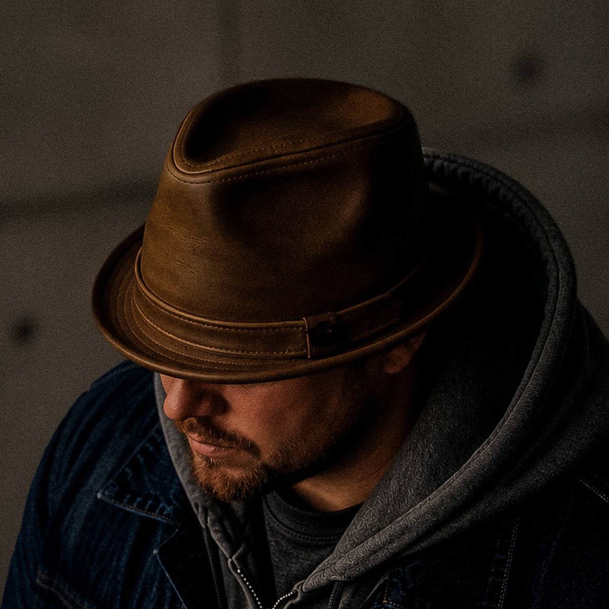 Man wearing the Noble Park brown structured leather fedora by Goorin Bros., handmade in the USA, paired with a gray hoodie and denim jacket, looking down; background is out of focus.