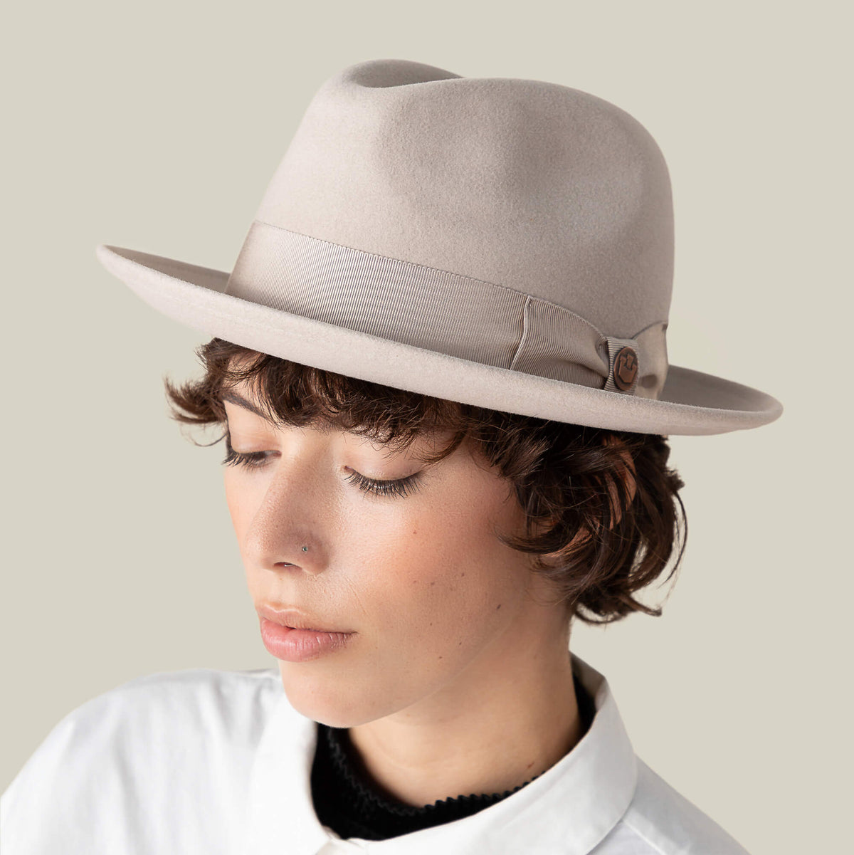 Person with short brown hair wearing "Dean the Butcher," a light beige fedora with grosgrain hatband by Goorin Bros., and a white collared shirt, looking down against a plain background.