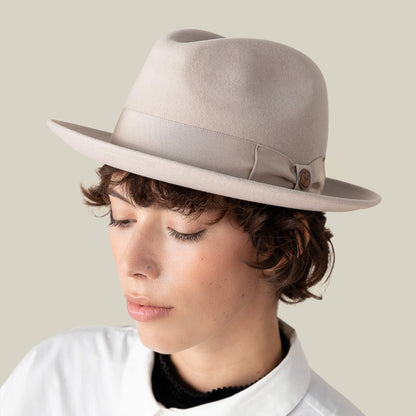 Person with short brown hair wearing "Dean the Butcher," a light beige fedora with grosgrain hatband by Goorin Bros., and a white collared shirt, looking down against a plain background.