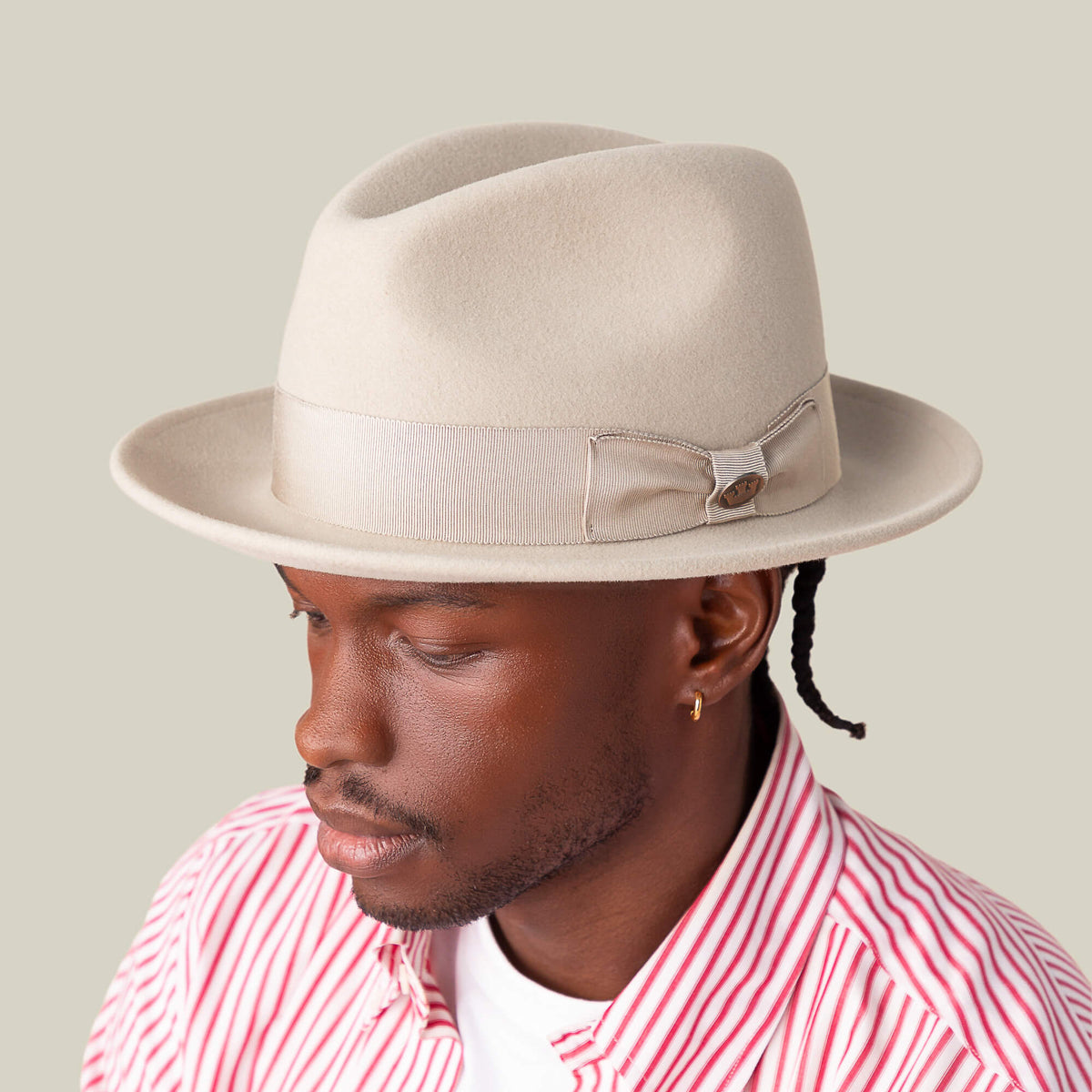 A man wearing the Goorin Bros. "Dean the Butcher" American-made fedora with grosgrain hatband and bow, paired with a pink and white striped shirt, looks down against a plain background.