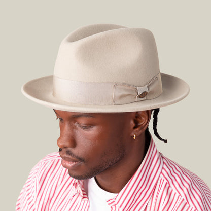 A man wearing the Goorin Bros. "Dean the Butcher" American-made fedora with grosgrain hatband and bow, paired with a pink and white striped shirt, looks down against a plain background.