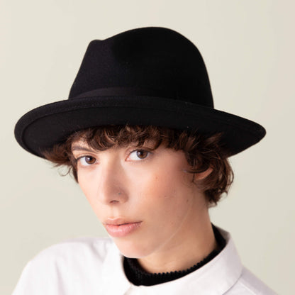 A person with short, curly brown hair wears the Goorin Bros. Dean the Butcher fedora—an American-made hat with a grosgrain band—paired with a white collared shirt, facing the camera against a plain light background.