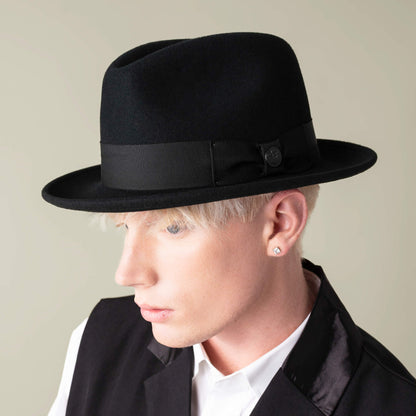 Person with short blond hair wears the Goorin Bros. "Dean the Butcher" American-made fedora with grosgrain hatband, black vest, and white shirt, looking slightly down and to the side against a neutral background.