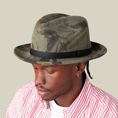 A man wearing Goorin Bros.' Dean the Butcher fedora with a green camo pattern and grosgrain hatband, a gold hoop earring, and a red and white striped shirt looks downward against a plain beige background.