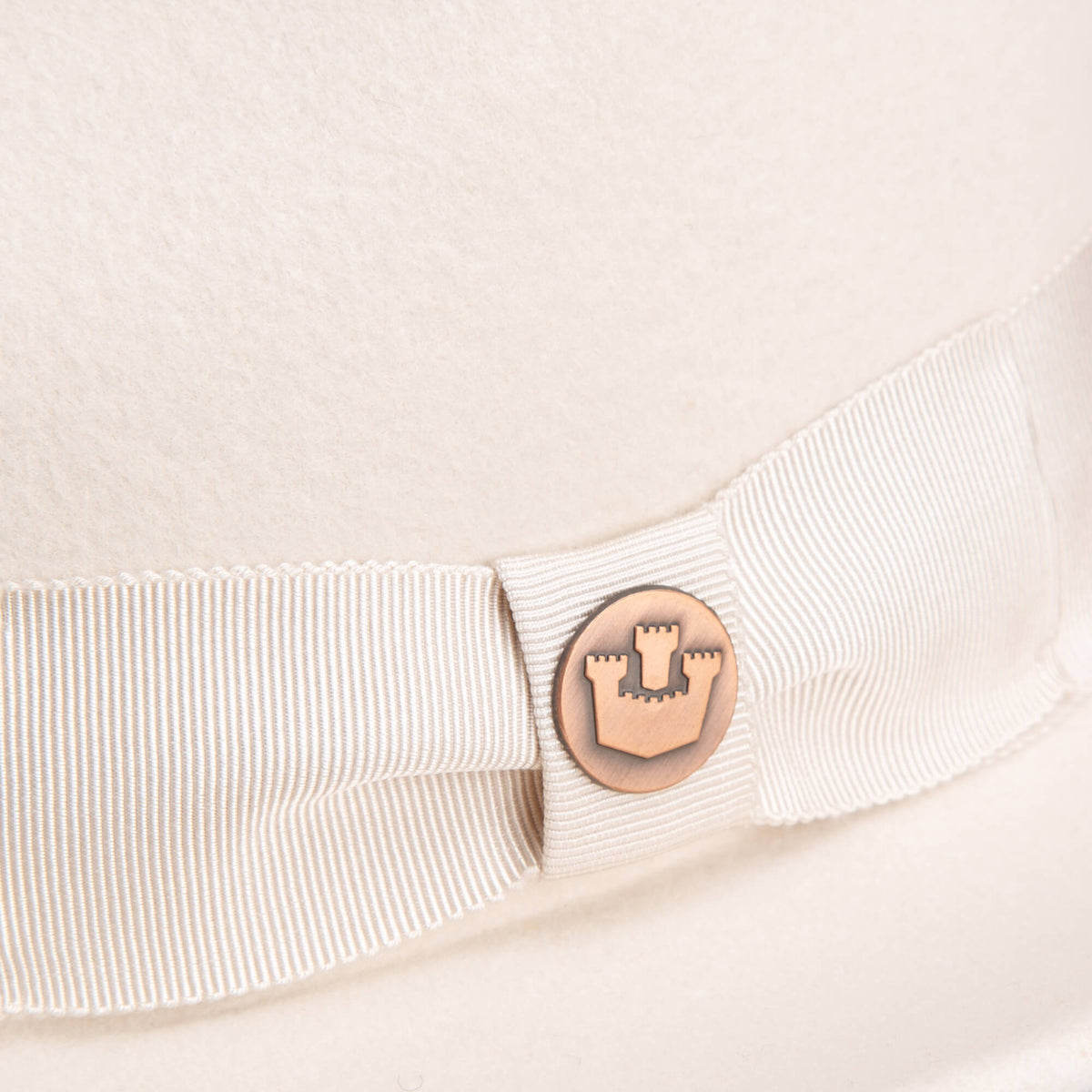 Close-up of the Goorin Bros. "Dean the Butcher" fedora in cream, featuring a grosgrain hatband and a round metal castle emblem.
