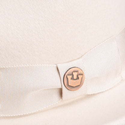 Close-up of the Goorin Bros. "Dean the Butcher" fedora in cream, featuring a grosgrain hatband and a round metal castle emblem.