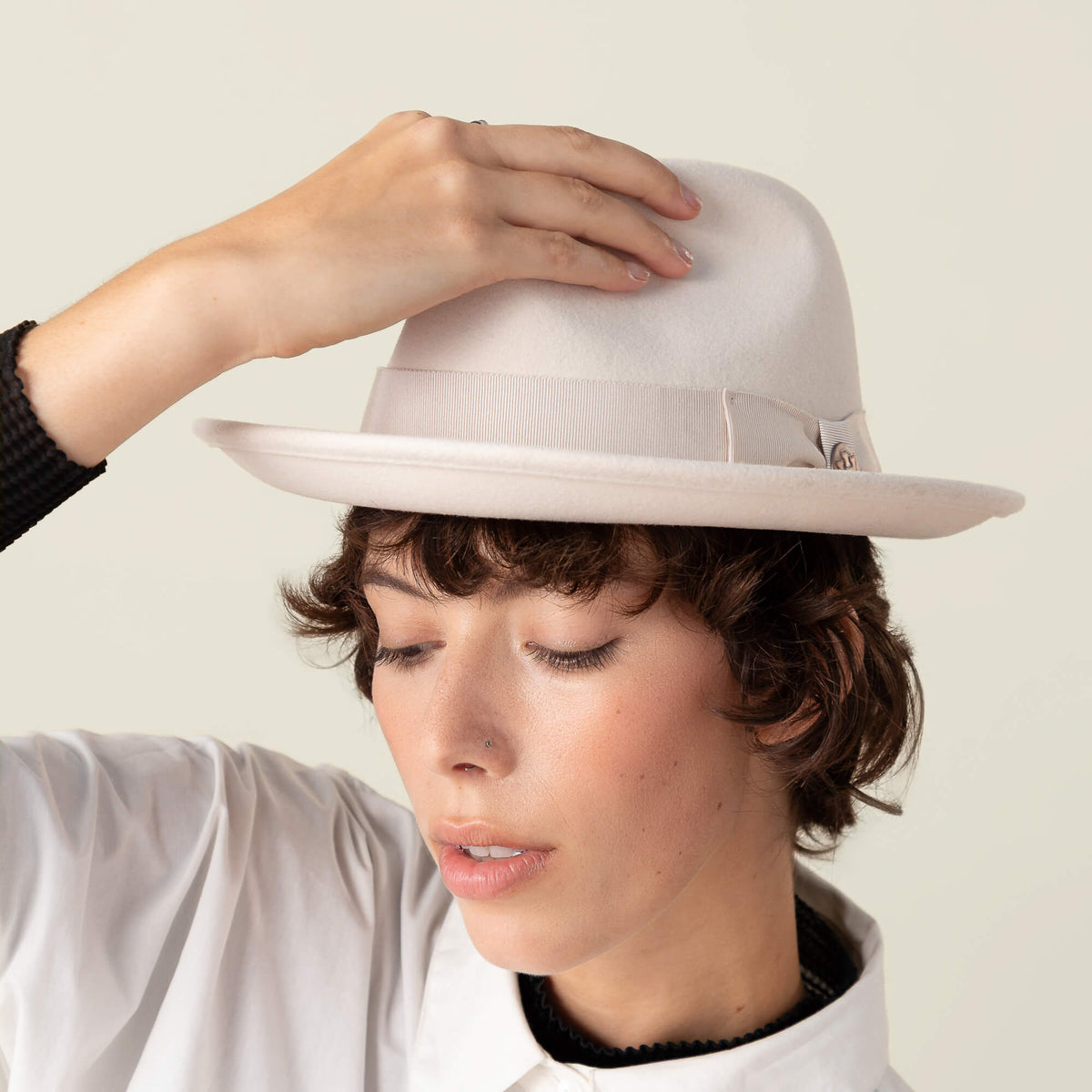 A person with short brown hair wears the Goorin Bros. Dean the Butcher hat, featuring a grosgrain hatband, and a white shirt, tilting the hat with one hand against a neutral background.