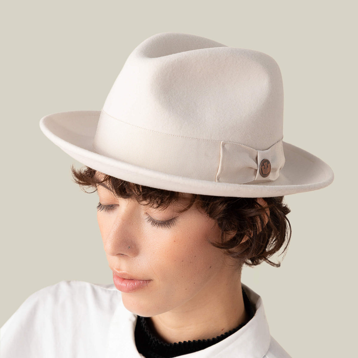 A person with short, curly brown hair wears Goorin Bros.’ Dean the Butcher fedora—American-made with grosgrain hatband and button detail—and a white collared shirt, looking downward against a plain background.