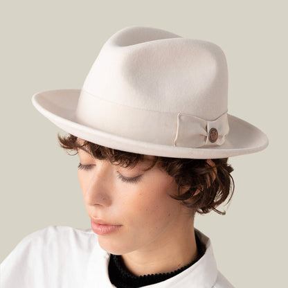 A person with short, curly brown hair wears Goorin Bros.’ Dean the Butcher fedora—American-made with grosgrain hatband and button detail—and a white collared shirt, looking downward against a plain background.