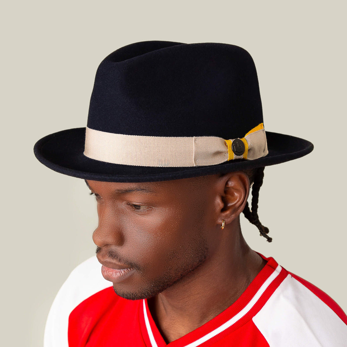 A man in the Dean the Butcher fedora by Goorin Bros., featuring a beige grosgrain hatband, red and white shirt, and small hoop earring, faces sideways against a plain light background.