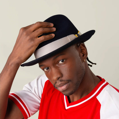 A man in a red and white shirt adjusts the Goorin Bros. "Dean the Butcher" fedora with a gray grosgrain band, looking directly at the camera against a plain light background.