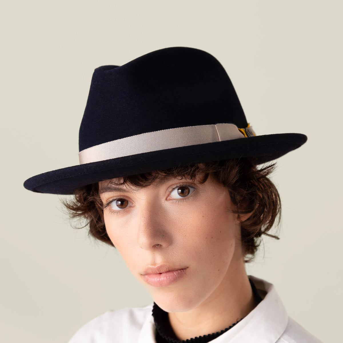 A person with short brown hair wears Dean the Butcher by Goorin Bros.—an American-made fedora with a grosgrain hatband—paired with a white shirt, gazing directly at the camera against a plain background.