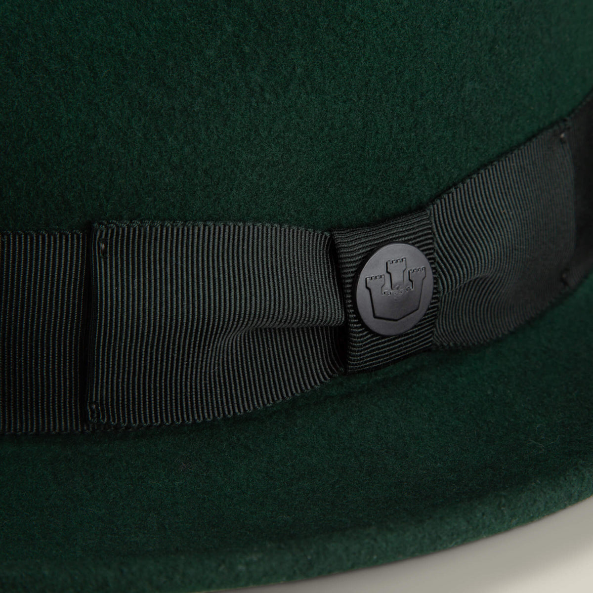 Close-up of the Dean the Butcher fedora by Goorin Bros., crafted in dark green felt with a black grosgrain hatband and a round metal castle emblem.