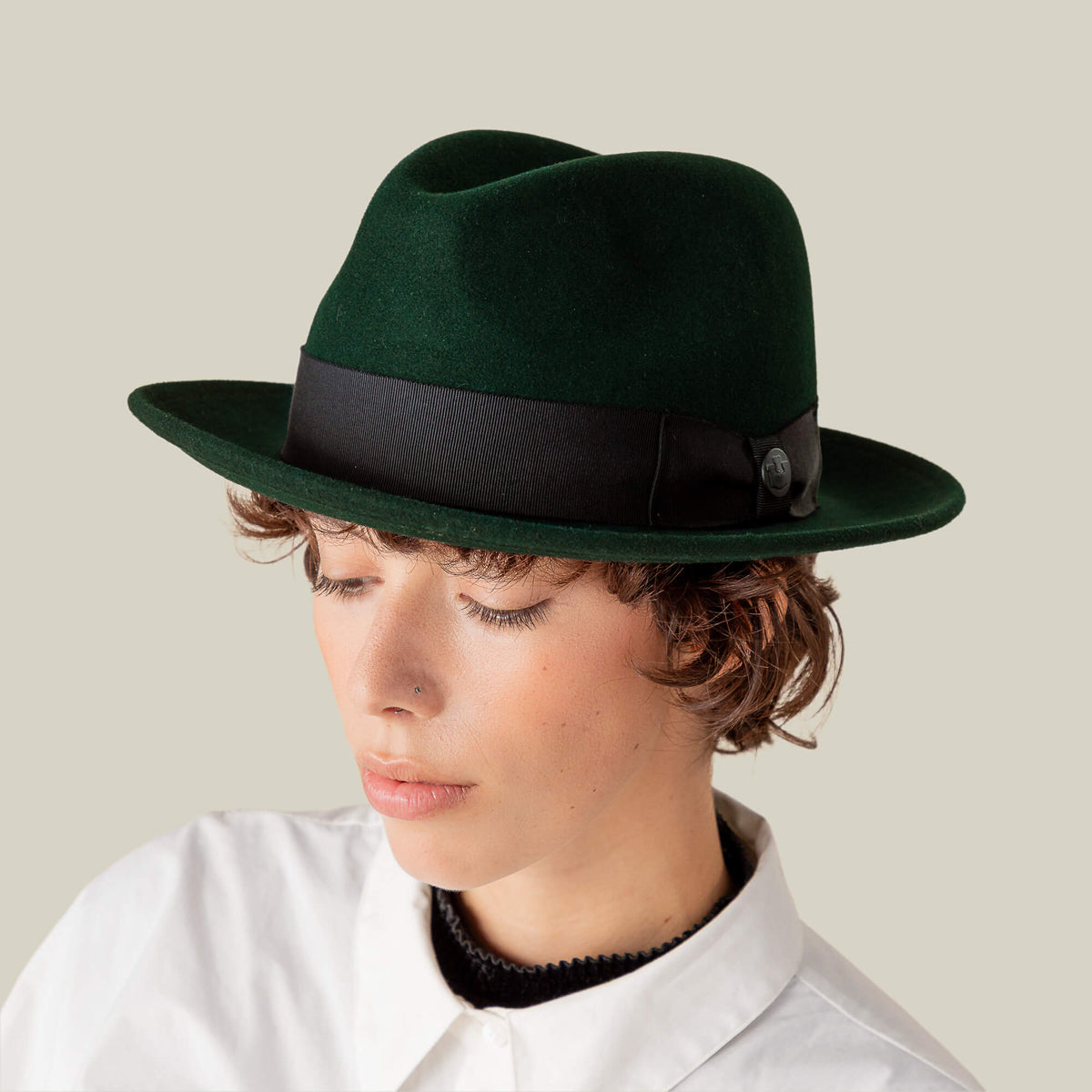 A person wearing the Dean the Butcher fedora by Goorin Bros., featuring a black grosgrain hatband, paired with a white button-up shirt and looking downward against a plain background.
