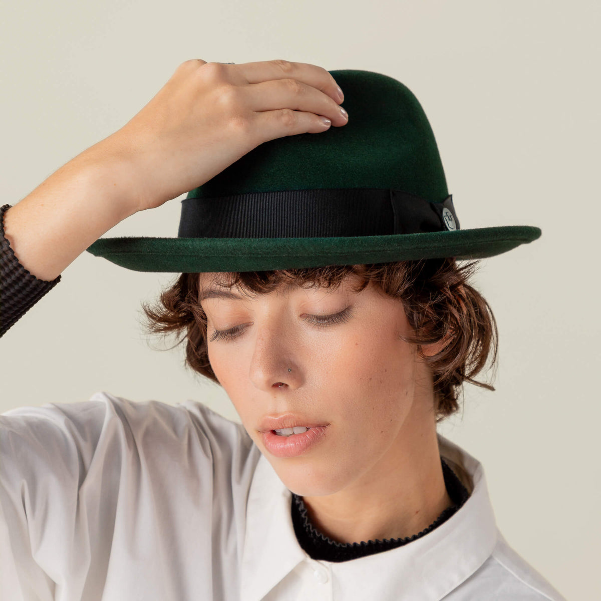 Person with short brown hair, eyes closed, wears a Goorin Bros. "Dean the Butcher" fedora with grosgrain hatband and white shirt, touching the top of the hat with one hand.