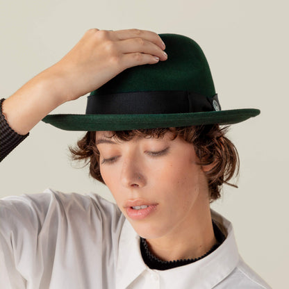 Person with short brown hair, eyes closed, wears a Goorin Bros. "Dean the Butcher" fedora with grosgrain hatband and white shirt, touching the top of the hat with one hand.