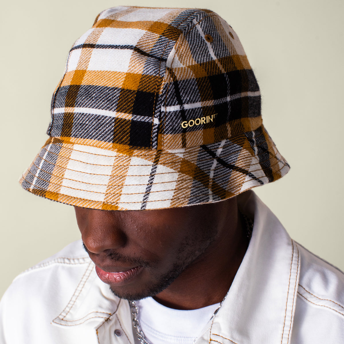 A person wears a white jacket and the Bespoke Wordmark Bucket by Goorin Bros., featuring a yellow, black, and white plaid design with an embroidered GOORIN wordmark on the side.