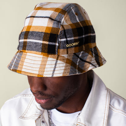 A person wears a white jacket and the Bespoke Wordmark Bucket by Goorin Bros., featuring a yellow, black, and white plaid design with an embroidered GOORIN wordmark on the side.