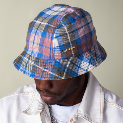 A person wearing the Bespoke Wordmark Bucket hat by Goorin Bros.—blue, pink, and white cotton linen with a GOORIN wordmark—dressed in a white jacket and looking downward.