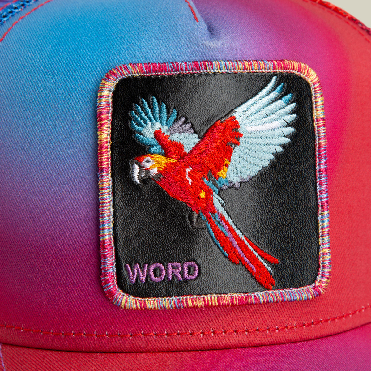 Close-up of The Farm’s “Bird is the Word” cap, showcasing a red parrot in flight and the word “WORD” stitched in purple on a black patch. All Sales Are Final.