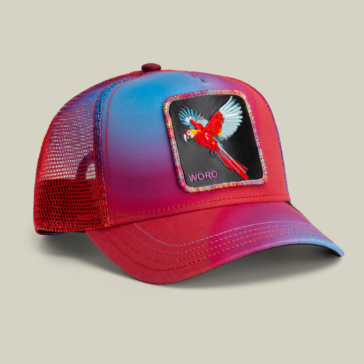 The Farm "Bird is the Word" mesh trucker hat in red and blue, featuring a flying parrot patch with "WORD" embroidered below. All sales are final.