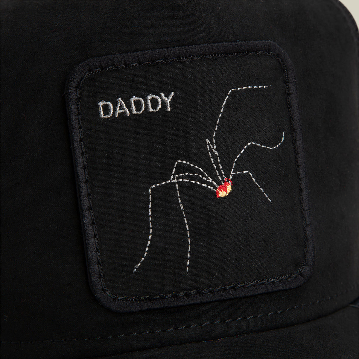A black fabric patch from Goorin Bros., named The Suede Spider, featuring the word “DADDY” and an embroidered arachnid with a red spot, is sewn onto a black surface.