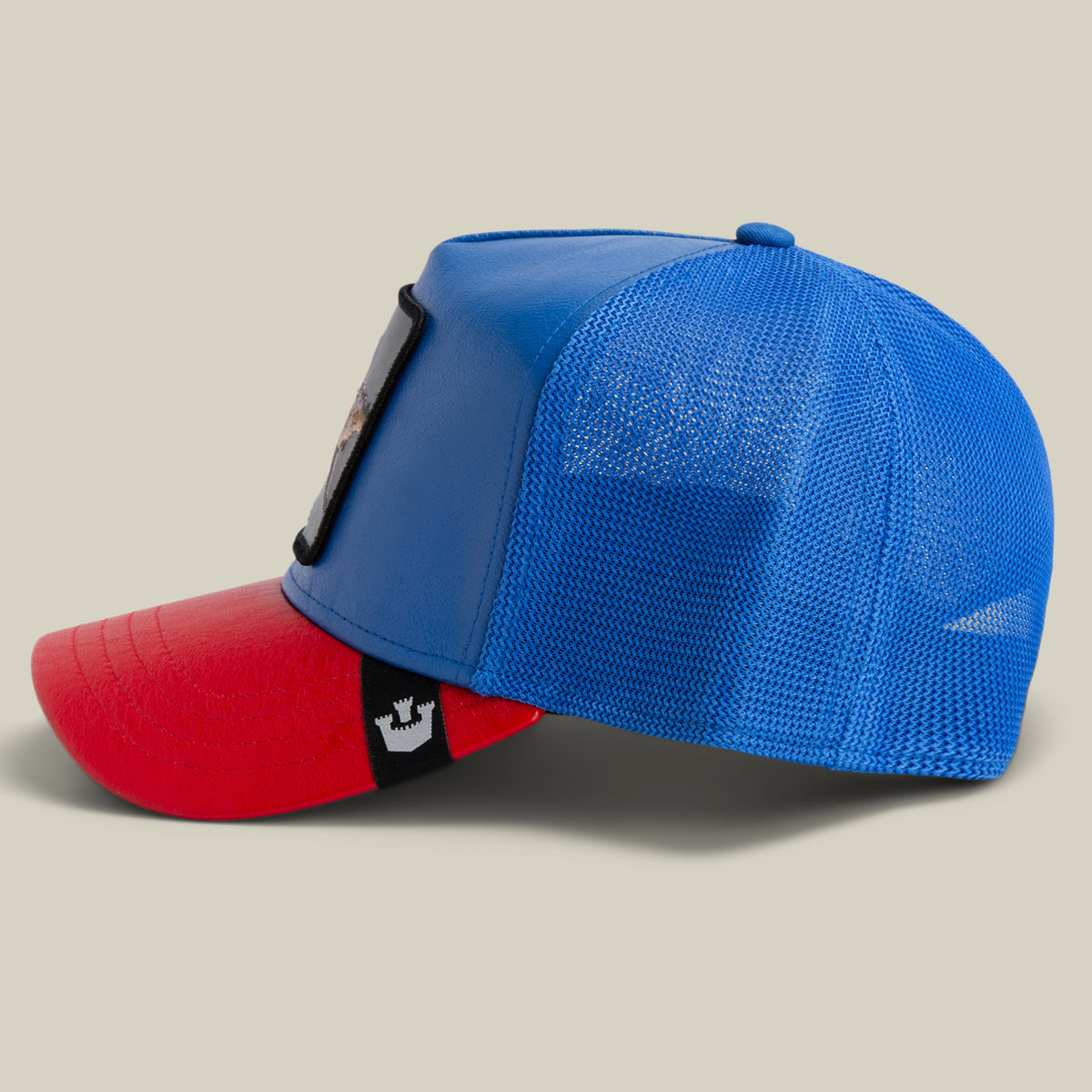 The Farm’s Deep Blue Fury trucker hat features blue and red panels, a mesh back, and a front patch. Displayed in side view on a plain background. All sales are final.