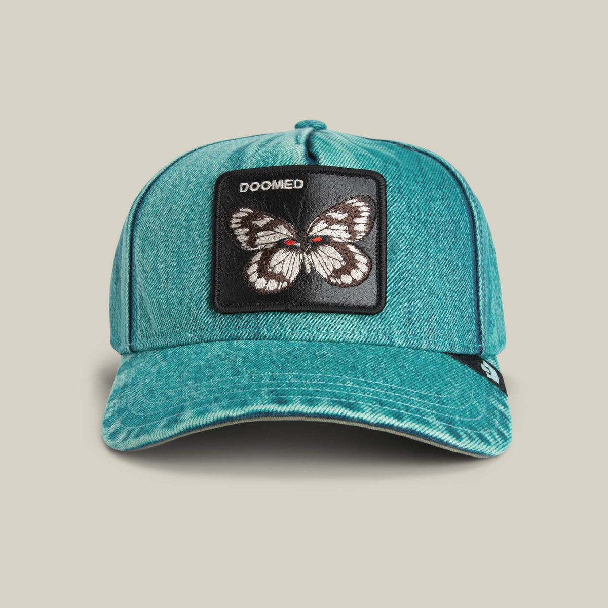 The Goorin Bros. Drawing Dead cap is a turquoise denim hat with thrift-store style, featuring a butterfly patch and "DOOMED" stitched above it.