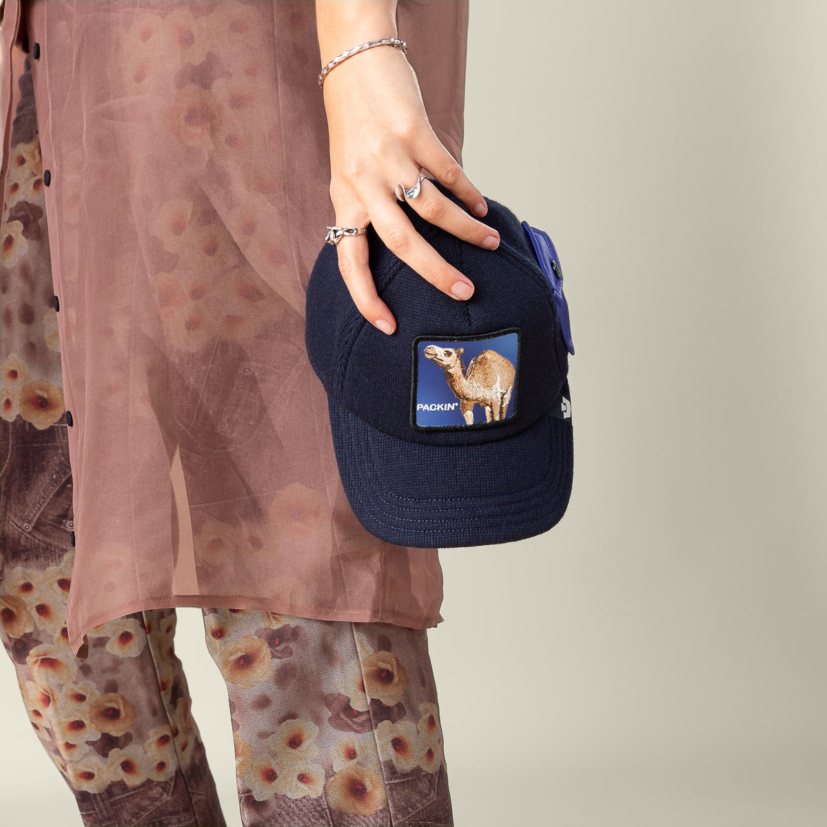 A person in floral pants and a sheer pink shirt holds the Goorin Bros. "Don't Say Fudge" navy blue cap with an animal patch, embracing ultimate comfort in their stylish look.