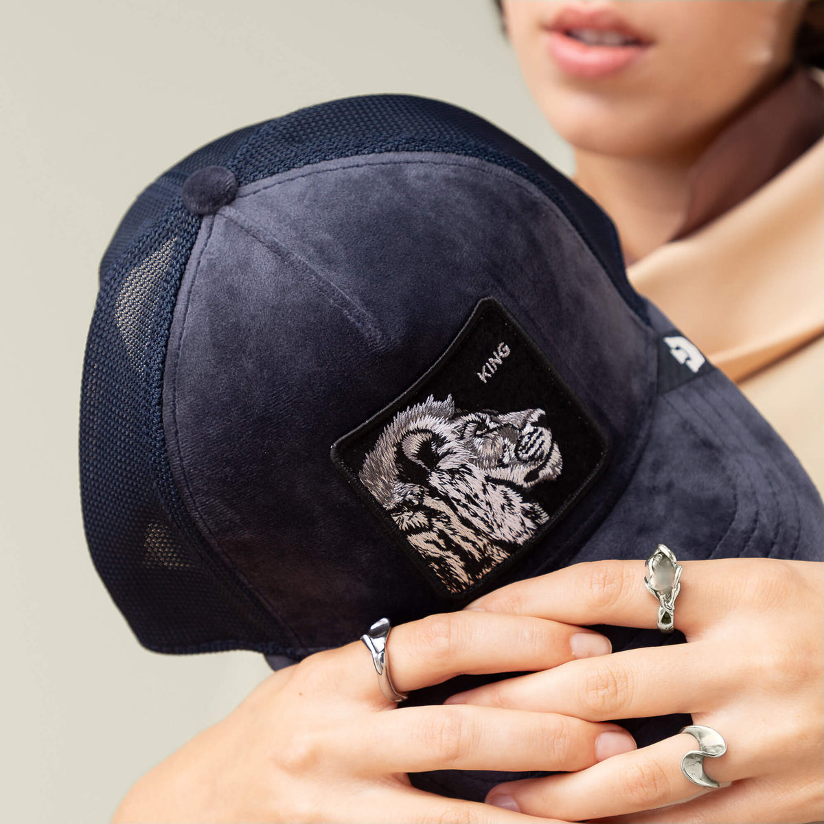 A person holds the Velour Lion cap by Goorin Bros.—a navy blue baseball cap featuring a KING patch and velour lion. They accessorize with silver rings and a light brown scarf.